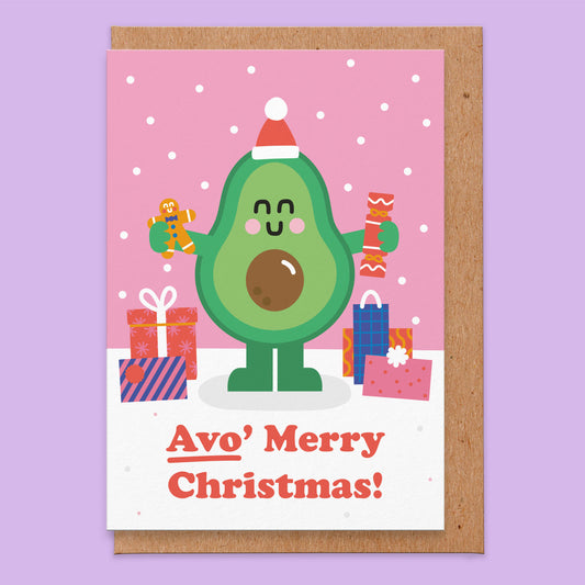 Greetings card that has an illustration of a Avocado holding a cracker and gingerbread man, wearing a Santa hat and presents on the ground and says Avo' Merry Christmas!