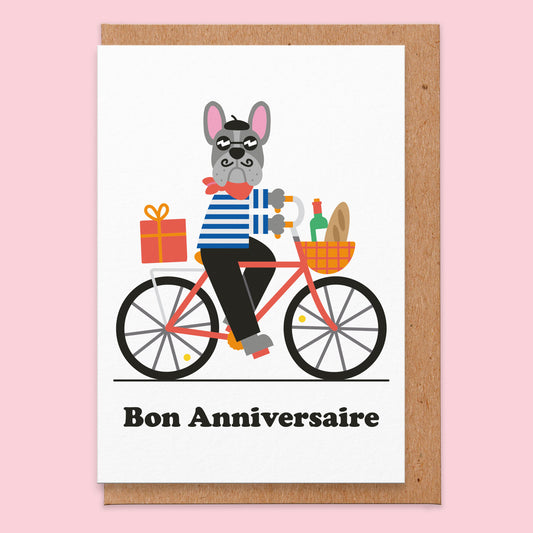Birthday card that reads bon anniversaire. The background is white and there is an illustration of a French bulldog riding a bicycle. In the bicycle basket there is a baguette and a bottle of red wine.