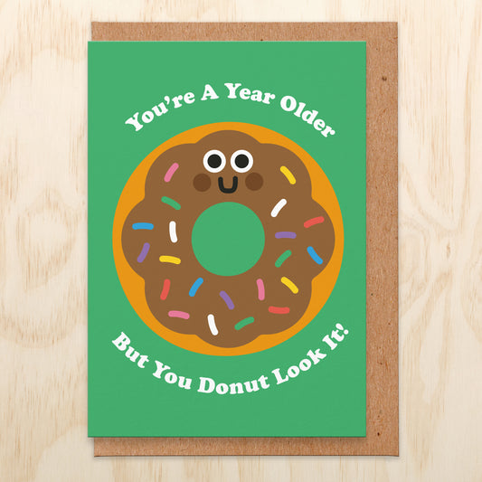 Birthday card that has a green background and an illustration of a chocolate donut with sprinkles on. The wording is you're a year older but you donut look it!