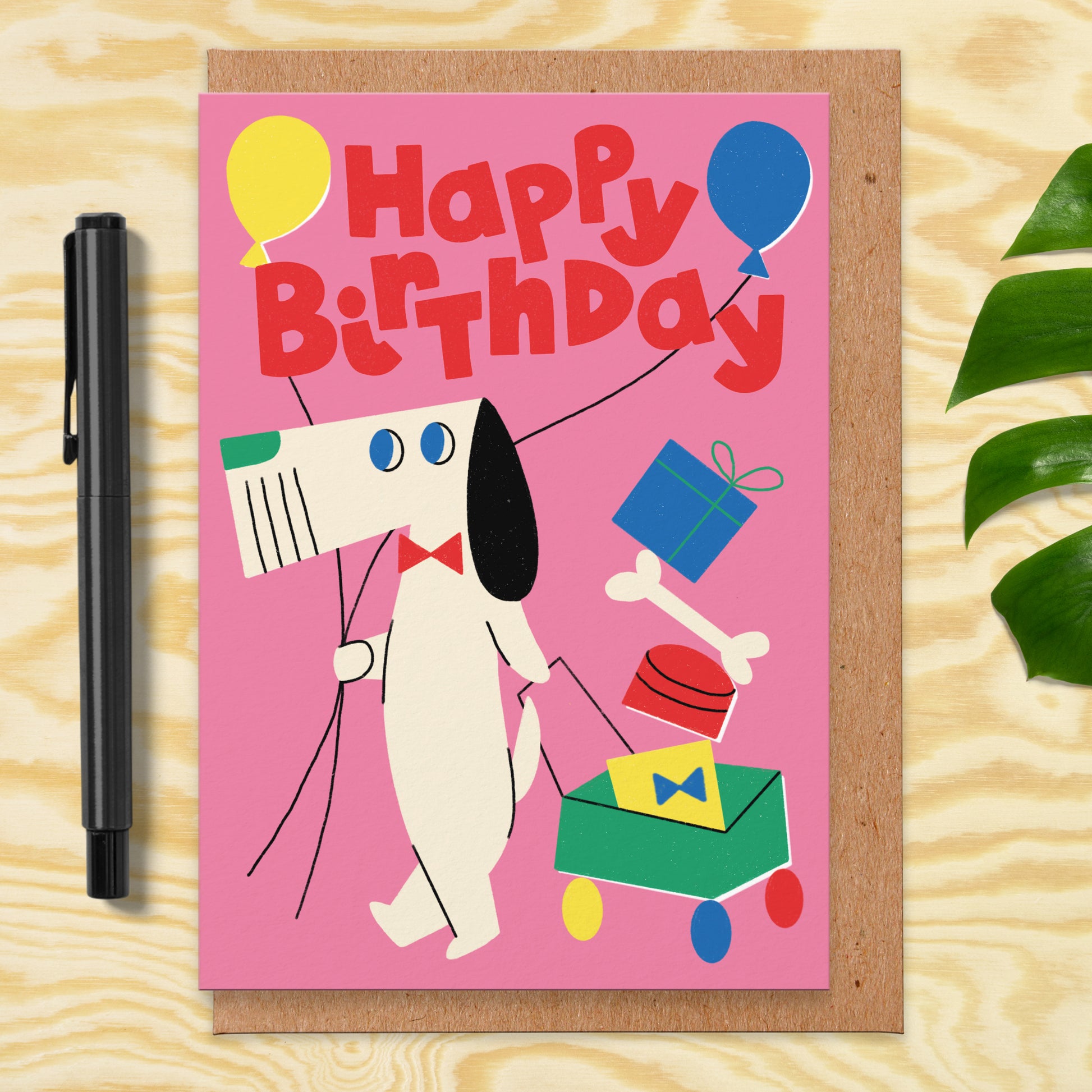 Pink greetings card that has an illustration of a dog holding balloons and pulling a trolley with presents in. The card reads happy birthday.